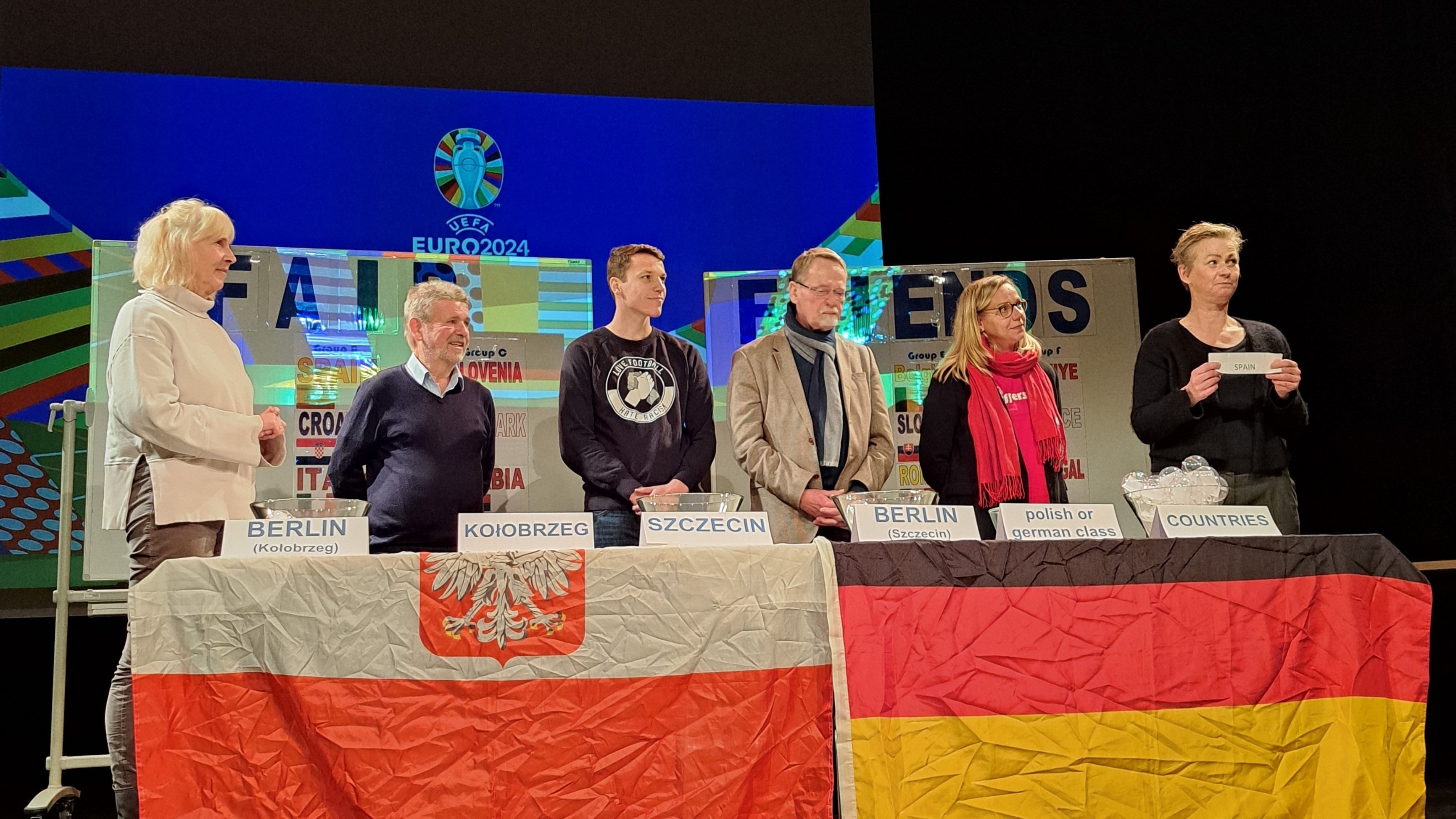 sechs Personen stehen hinter einem Tisch mit Schildern drauf und der polnischen und deutschen Flagge davor